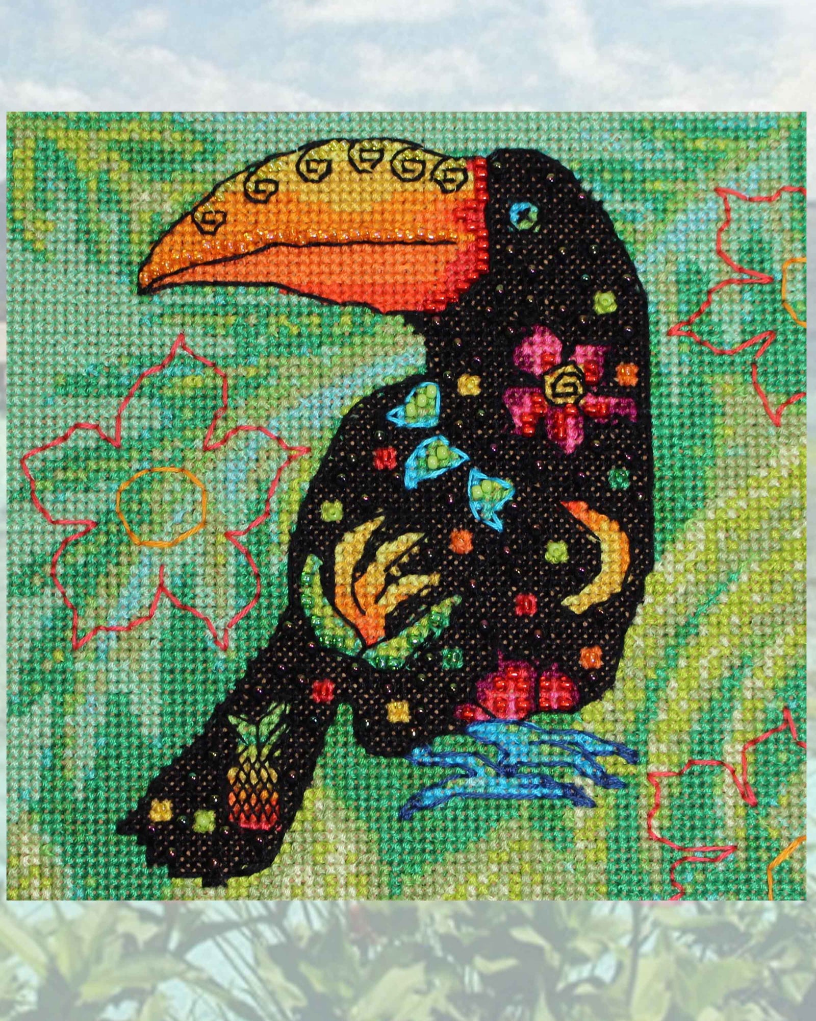 Fruity Toucan Cross Stitch Pattern completed design features an large-billed black toucan with pink flowers, green leaves, yellow bananas and pineapple decorated body with colorful spots on a leafy green background.  Buy purchase for sale PDF Pattern available at Raspberry Lane Crafts.